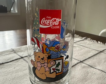 May include: Clear Coca-Cola Classic glass featuring a Christmas-themed design. The glass has a red Coca-Cola logo and illustrations of a teddy bear, a clown, a Christmas tree, and other festive elements.