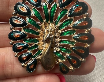 May include: A vintage peacock brooch with a gold-tone metal frame. The brooch features a detailed peacock design with green, black, and orange enamel accents. The peacock's tail feathers are spread out, creating a circular shape.