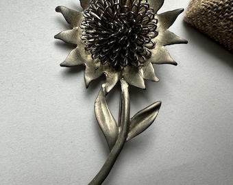 May include: A vintage silver-toned metal sunflower brooch. The brooch features a detailed flower head with a wire center and pointed petals. The stem has two leaves. A classic floral accessory.