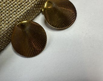 May include: Two gold-toned, round clip-on earrings. Each earring features a fan-like design with radiating lines. The earrings are set against a neutral background with a textured, woven fabric.
