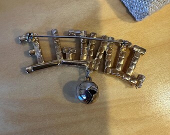 May include: A gold-toned brooch spelling out the word "JUNE" in a stylized font, embellished with clear rhinestones. A small, silver-colored globe charm dangles from the bottom. The brooch is set against a light brown wooden surface.