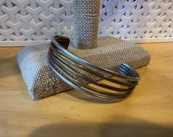 May include: Silver-toned cuff bracelet with five parallel bands. The bracelet is displayed on a burlap-covered jewelry stand against a white lattice background. The cuff is a simple, elegant design.