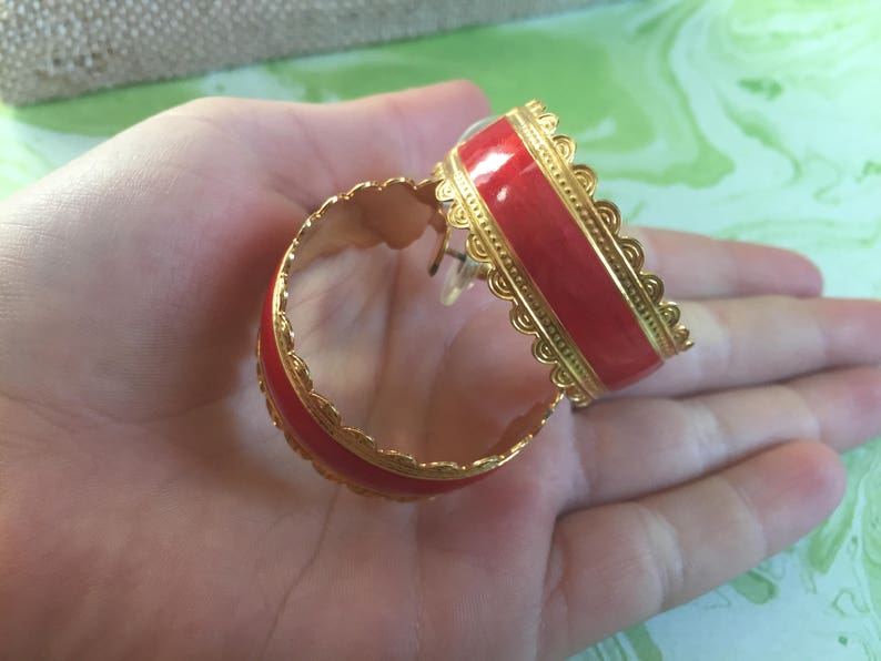 May include: A pair of gold hoop earrings with a red enamel inlay. The earrings have a decorative, scalloped edge.