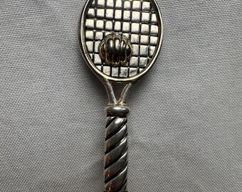May include: Silver-toned tennis racket pendant. The racket head features a grid pattern, with a decorative element at the center. The handle is detailed with a twisted rope design. A sports-themed accessory.