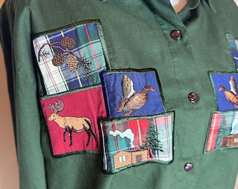 May include: A green button-up shirt with embroidered patches featuring a moose, a duck, pine cones, and a cabin. The patches are on a plaid background.