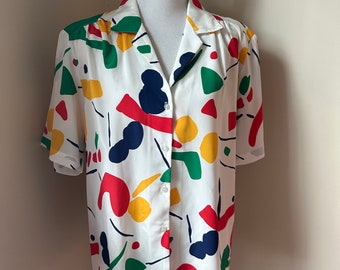 May include: White button-down shirt with a colorful abstract pattern. The short-sleeved shirt features a collared neckline and is adorned with red, yellow, green, and navy blue shapes. The shirt is displayed on a mannequin.