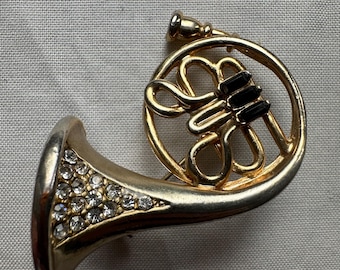 May include: A gold-toned brooch in the shape of a French horn. The bell of the horn is encrusted with clear rhinestones, and the body features black accents. The brooch is a musical instrument-themed accessory.