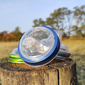 Op de afbeelding: Een close-up van een doorzichtig plastic deksel met een blauwe en groene rand. Op het deksel staat "Ruby" gedrukt. Het deksel ligt op een houten paal.