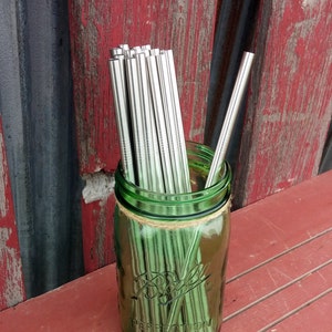 Puede incluir: Un tarro de vidrio verde con una cuerda marrón atada alrededor de la parte superior, lleno de un paquete de pajitas para beber de metal plateado.