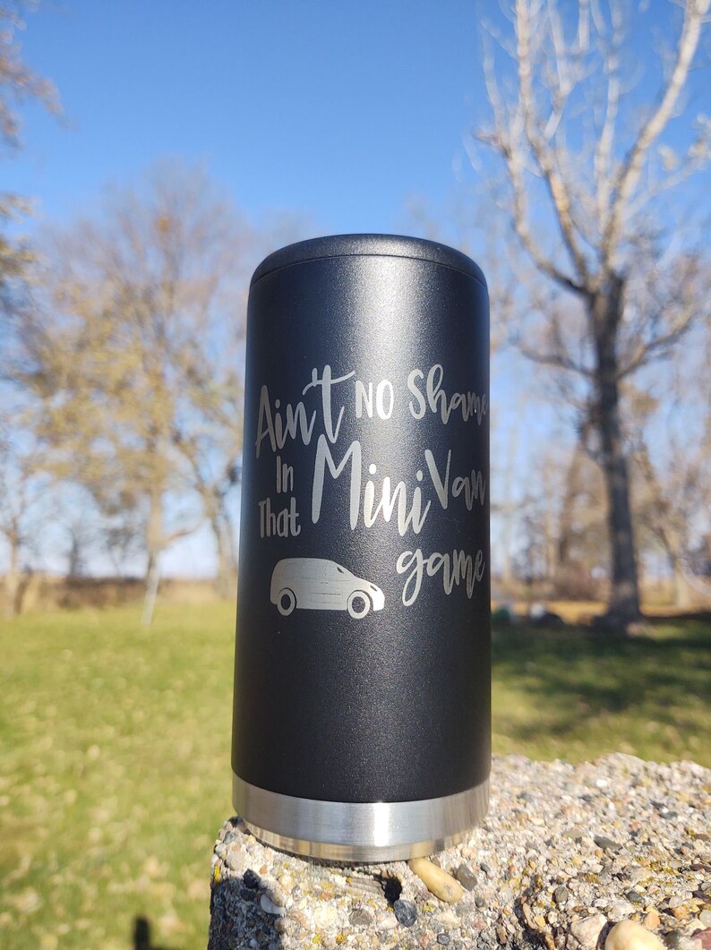 May include: A black stainless steel can cooler with a silver band around the top and bottom. The cooler has an engraved design that says "Ain't no shame in that Mini Van game" with a silhouette of a minivan.