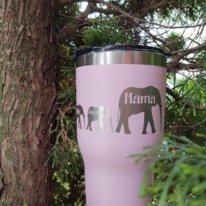 Puede incluir: Un vaso t&eacute;rmico rosa con tapa negra y borde plateado. El vaso presenta una familia de elefantes con la palabra "Mama" y el nombre "James" grabado en el lateral. El vaso est&aacute; sobre un fondo natural.