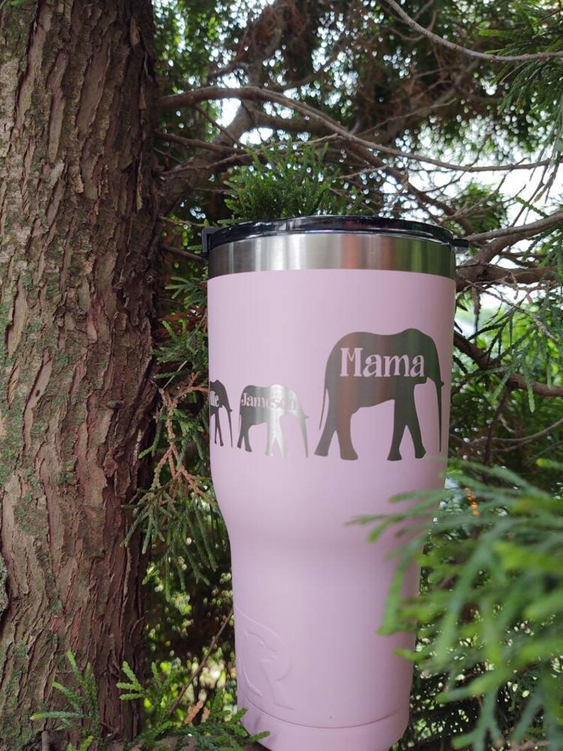 Puede incluir: Un vaso t&eacute;rmico rosa con tapa negra y borde plateado. El vaso presenta una familia de elefantes con la palabra "Mama" y el nombre "James" grabado en el lateral. El vaso est&aacute; sobre un fondo natural.