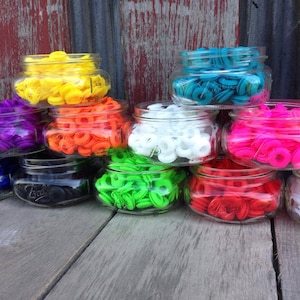 May include: Assortment of colorful plastic grommets in glass jars. The grommets are in various colors, including yellow, orange, purple, blue, green, white, pink, and red.