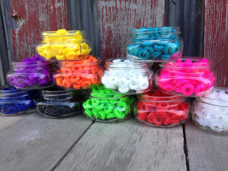May include: Assortment of colorful plastic grommets in glass jars. The grommets are in various colors, including yellow, orange, purple, blue, green, white, pink, and red.