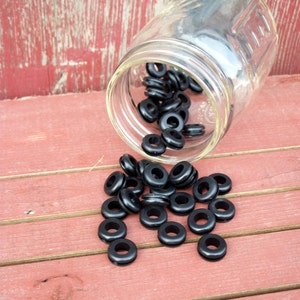 Op de afbeelding: Een heldere glazen pot die is omgekieperd en zwarte rubberen grommets op een verweerd rood houten oppervlak laat vallen. De grommets zijn cirkelvormig met een gat in het midden.