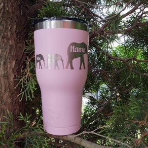 Puede incluir: Un vaso t&eacute;rmico rosa con tapa negra y borde plateado. El vaso presenta una familia de elefantes con las palabras "Allie", "Emerson" y "Mama" grabadas en el lateral. El vaso est&aacute; sobre una rama de &aacute;rbol.