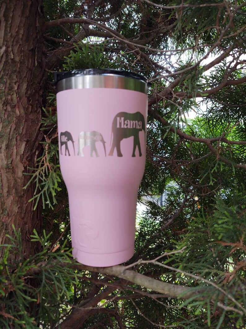 Puede incluir: Un vaso t&eacute;rmico rosa con tapa negra y borde plateado. El vaso presenta una familia de elefantes con las palabras "Allie", "Emerson" y "Mama" grabadas en el lateral. El vaso est&aacute; sobre una rama de &aacute;rbol.