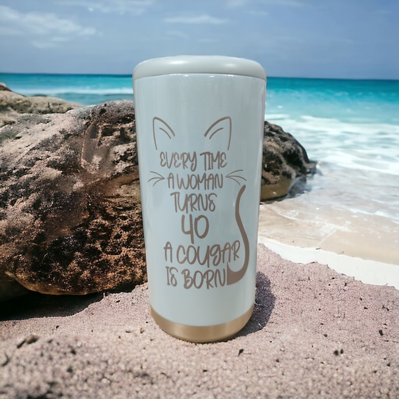 May include: A white stainless steel tumbler with a gold lid and a cat silhouette. The tumbler has the text "Every time a woman turns 40 a cougar is born" printed on it.