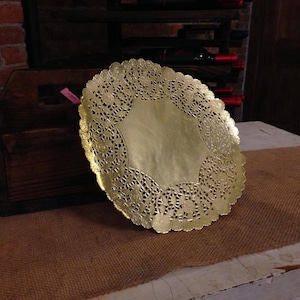 May include: A gold-coloured paper doily with a delicate lace pattern. The doily is round and has a centre hole. It is sitting on a brown hessian surface.