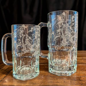 Op de afbeelding: Twee doorzichtige glazen bierpullen van verschillende groottes, elk voorzien van een gedetailleerde witte ets van een gevleugelde draak. De pullen hebben stevige handvatten en een gefacetteerde voet, wat het drakenontwerp op het glas toont.