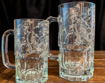 May include: Two clear glass beer mugs of different sizes, each featuring a detailed white etched graphic of a winged dragon. The mugs have sturdy handles and a faceted base design, showcasing the intricate dragon artwork on the transparent glass.