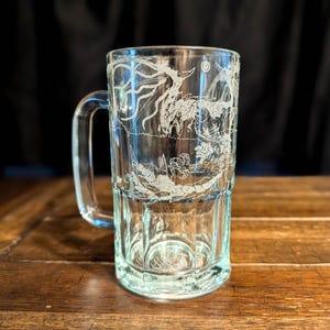 Può includere: Un boccale da birra in vetro trasparente con un'incisione bianca dettagliata di un drago con ali spiegate e una lunga coda. Il boccale presenta un manico robusto e una base spessa con pannelli verticali nella parte inferiore del vetro.