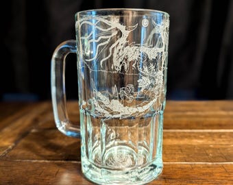 May include: A clear glass beer mug featuring a white etched design of a dragon with wings spread and a long tail. The mug has a sturdy handle and a thick base with vertical panels. The dragon illustration wraps around the exterior surface.