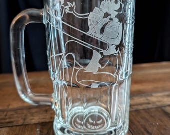 May include: A clear glass beer mug featuring an etched design of a character sitting at a counter while holding a drink. The base of the mug displays an etched graphic of two circular faces with wide, toothy grins and triangular eyes.