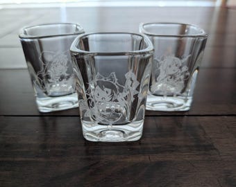 May include: Three clear glass shot glasses with square bases and rounded rims. Each glass features a white etched graphic of an anime-style character with large eyes and hair accessories, positioned in the center of the front-facing glass surface.