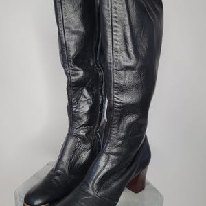 May include: A pair of black knee-high boots with a slight sheen. The boots have a rounded toe with a gold-colored trim, a block heel with a dark woodgrain finish, and a tall shaft. The boots are made of a smooth material.