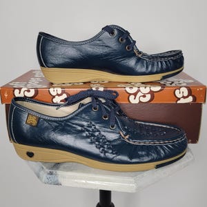 May include: A pair of navy blue leather shoes with tan soles and laces. The shoes are displayed on top of a brown and orange box. The shoes have a woven design on the front.