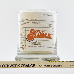 Peut inclure: Un bocal en verre pour bougie avec une &eacute;tiquette qui indique "Clove + Orange" et "Banned Books Collection". L'&eacute;tiquette est une r&eacute;plique d'une carte de biblioth&egrave;que avec une date d'&eacute;ch&eacute;ance du livre du 9 juin 1962. Le bocal &agrave; bougie est pos&eacute; sur un livre intitul&eacute; "A Clockwork Orange" d'Anthony Burgess.