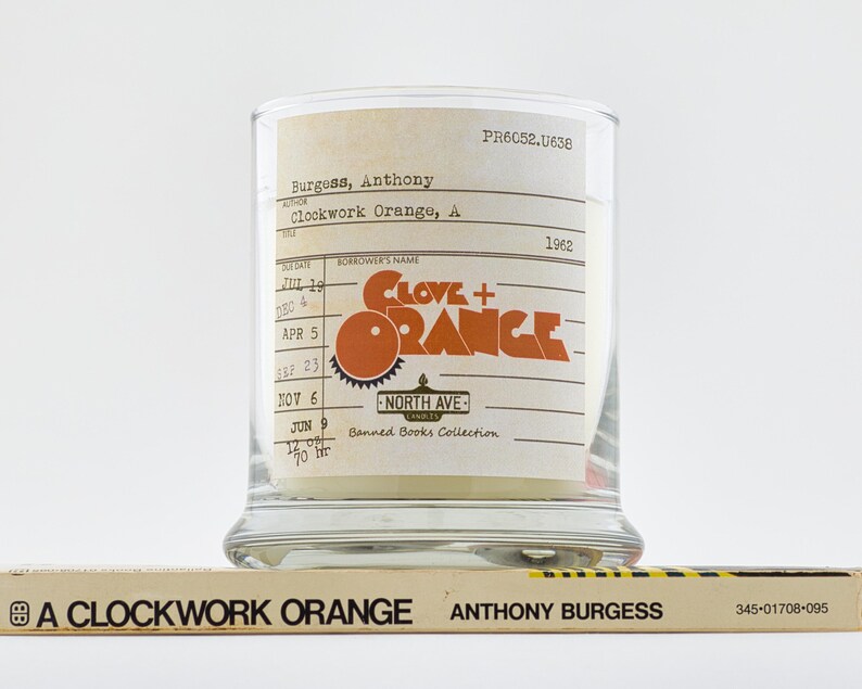 Peut inclure: Un bocal en verre pour bougie avec une &eacute;tiquette qui indique "Clove + Orange" et "Banned Books Collection". L'&eacute;tiquette est une r&eacute;plique d'une carte de biblioth&egrave;que avec une date d'&eacute;ch&eacute;ance du livre du 9 juin 1962. Le bocal &agrave; bougie est pos&eacute; sur un livre intitul&eacute; "A Clockwork Orange" d'Anthony Burgess.