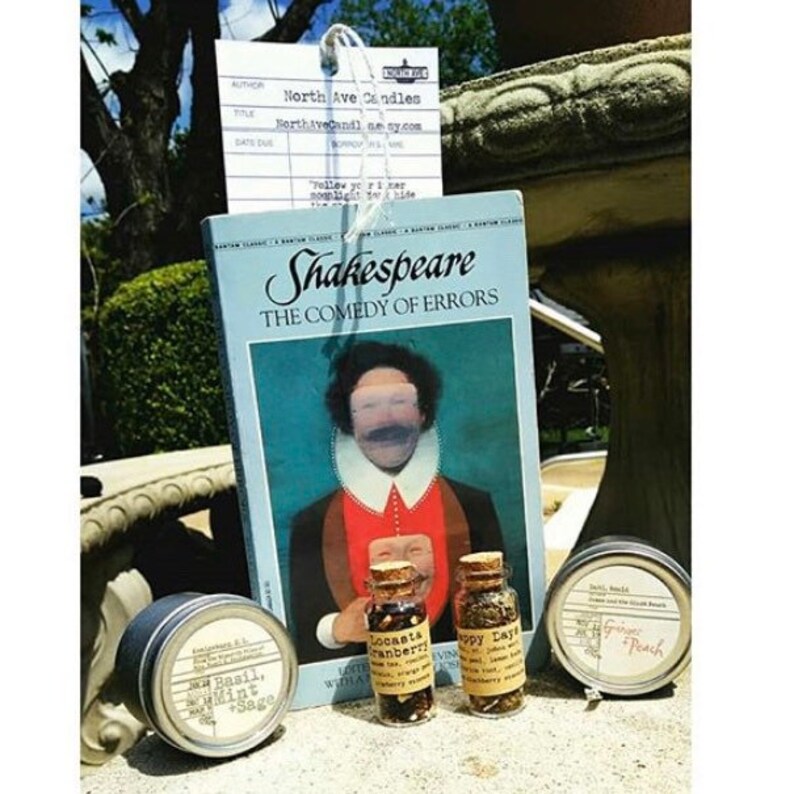 May include: A collection of small glass jars filled with colorful herbs and spices. The jars are labeled with names like "Cranberry", "Happy Days", and "Ginger Peach". The jars are sitting on a table next to a copy of Shakespeare's "The Comedy of Errors".