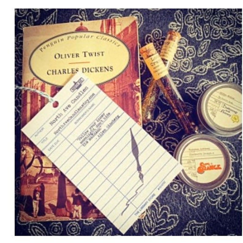 May include: A vintage Penguin Classics edition of Oliver Twist by Charles Dickens, a book with a worn cover, is displayed on a dark patterned surface with a small, white, lined card, a small glass vial, and two small round containers. The card has the text "North Ave Candles" and "northavecandles.etsy.com".