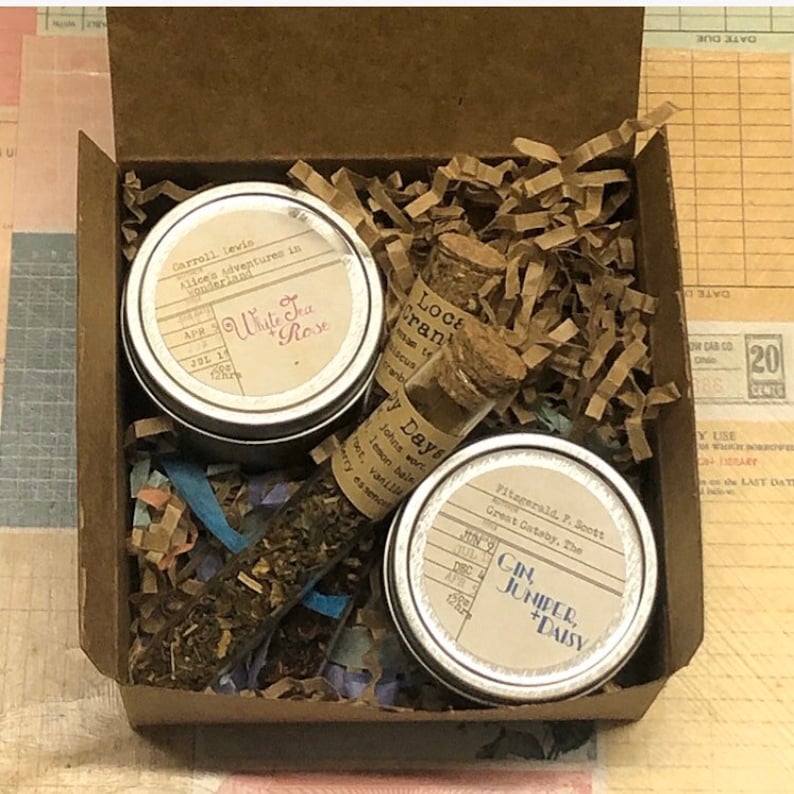 May include: A brown cardboard box filled with brown packing paper and three small metal tins. The tins are labeled with book titles and authors, including "White Tea + Rose", "Local Cranberry", and "Gin, Juniper, + Daisy".