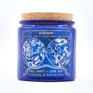 May include: A blue glass candle jar with a cork lid. The jar has a white label with the text "ASTRONOMY. the scientific study of celestial objects and phenomena PALO SANTO + STAR DUST Aromachology by North Ave Candles" and a white illustration of a constellation map.