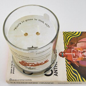 Peut inclure: Un bocal &agrave; bougie en verre transparent avec une &eacute;tiquette blanche qui dit "What's it gonna be then, eh?" Le bocal &agrave; bougie est pos&eacute; sur un livre intitul&eacute; "A Clockwork Orange" d'Anthony Burgess. La couverture du livre pr&eacute;sente une illustration color&eacute;e d'une moto.