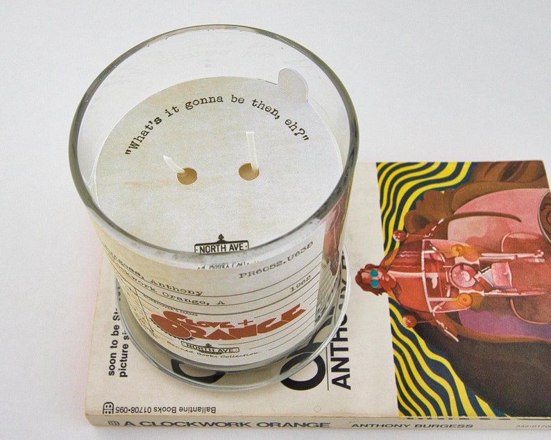 Peut inclure: Un bocal &agrave; bougie en verre transparent avec une &eacute;tiquette blanche qui dit "What's it gonna be then, eh?" Le bocal &agrave; bougie est pos&eacute; sur un livre intitul&eacute; "A Clockwork Orange" d'Anthony Burgess. La couverture du livre pr&eacute;sente une illustration color&eacute;e d'une moto.