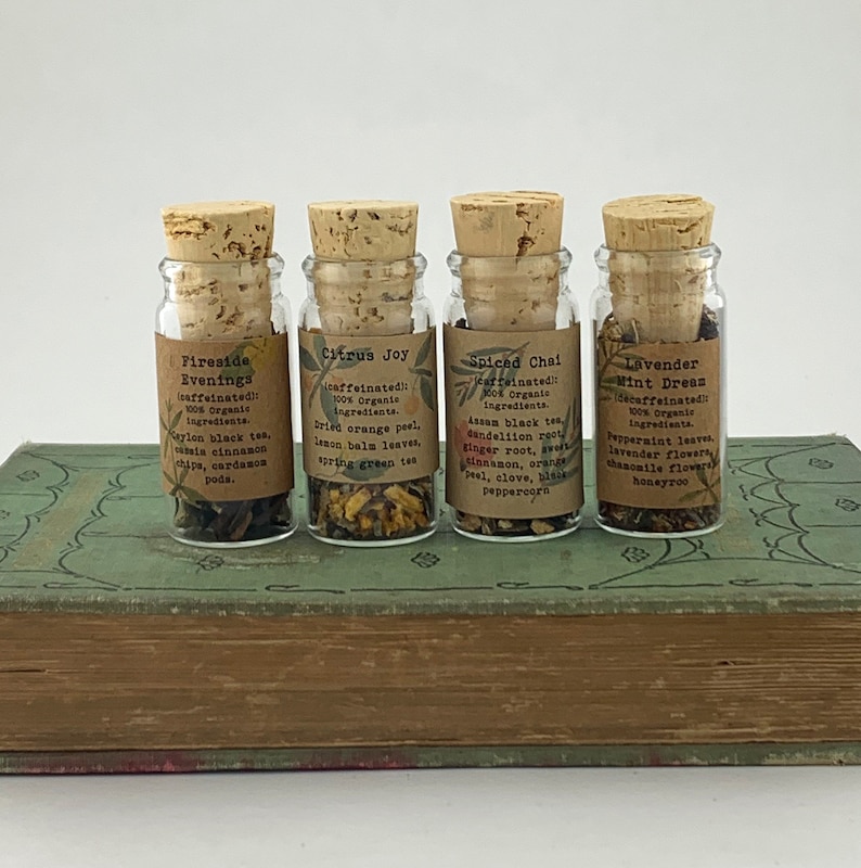 May include: Four small glass bottles with cork stoppers, each filled with loose leaf tea blends. The labels are printed with the names of the tea blends: Fireside Evenings, Citrus Joy, Spiced Chai, and Lavender Mint Dream. The bottles are sitting on a closed book.