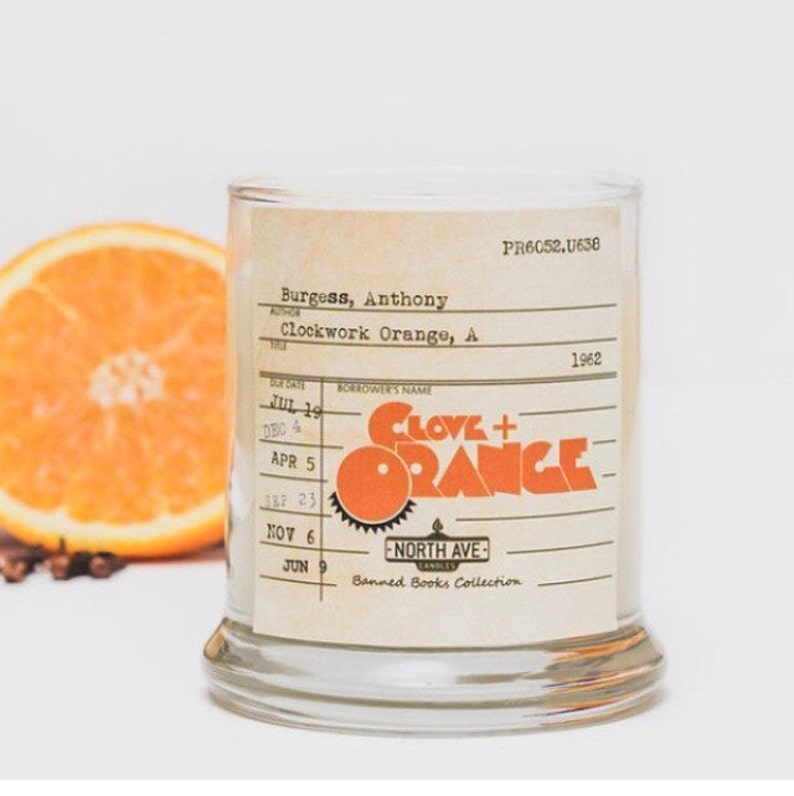 Peut inclure: Un bocal en verre pour bougie avec une &eacute;tiquette qui indique "Clove + Orange" et "Banned Books Collection". L'&eacute;tiquette est stylis&eacute;e comme une carte de biblioth&egrave;que avec le titre du livre "Clockwork Orange" d'Anthony Burgess.