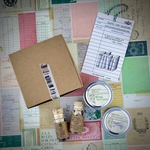 May include: A brown box with a book-themed washi tape on the side, two small glass bottles with labels that say "Lavender Mint Dress" and "Petit Grain", and two metal tins with labels that say "The Shire Sweetgrass" and "Austen Jane Petit Grain Perfume". The image also includes a library card with the text "A book is a dress you hold in your hand - Neil Gaiman" and the title "North Ave Candles".
