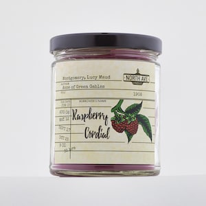 May include: A glass jar candle with a pink label that says "Raspberry Cordial" and features a vintage library card design. The label also includes the text "Montgomery, Lucy Maud" and "Anne of Green Gables". The jar has a black lid and is labeled "North Ave Candles".