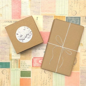 May include: Two brown gift boxes wrapped in kraft paper with white twine. One box has a round sticker with a black and white illustration of a ship with birds flying above it. The other box has a simple bow.
