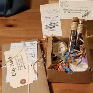 May include: A brown box filled with a book, candles, and tea. The book is wrapped in brown paper and tied with twine. The candles are in small glass jars and the tea is in glass tubes. The box is filled with shredded paper and has a label that says "City Books".