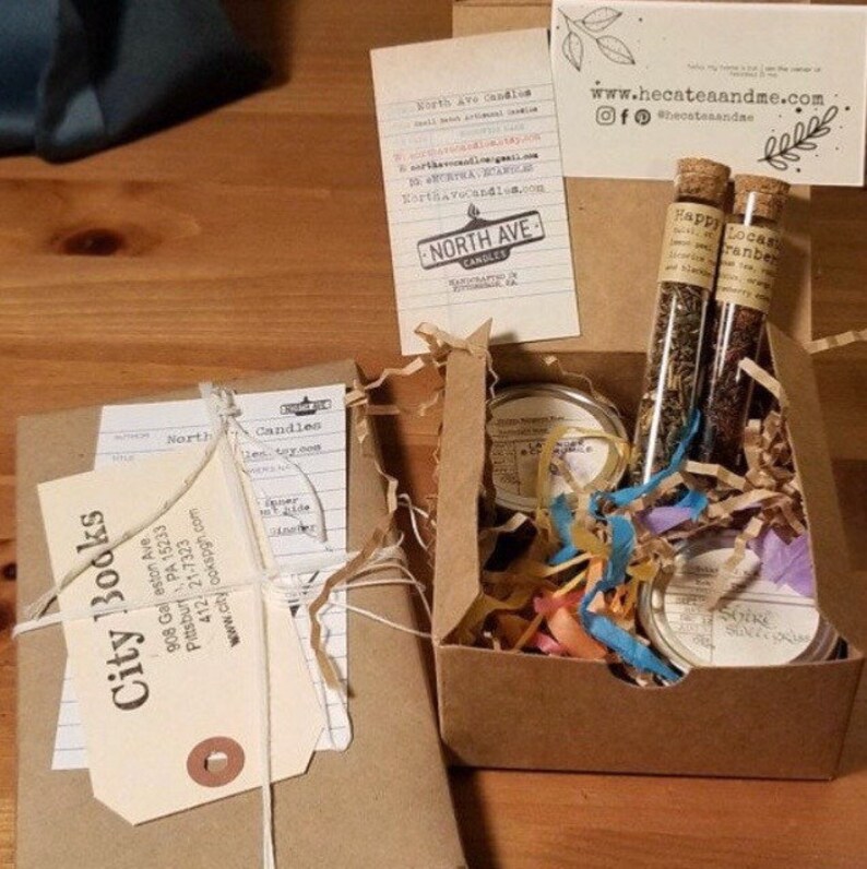 May include: A brown box filled with a book, candles, and tea. The book is wrapped in brown paper and tied with twine. The candles are in small glass jars and the tea is in glass tubes. The box is filled with shredded paper and has a label that says "City Books".