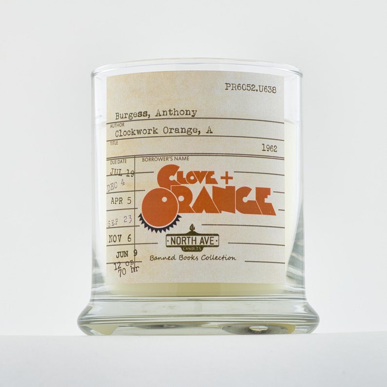 Peut inclure: Une bougie en verre avec une &eacute;tiquette blanche sur laquelle est inscrit "Clove + Orange" dans une police r&eacute;tro. L'&eacute;tiquette comprend &eacute;galement le texte "Banned Books Collection" et "North Ave Candles".