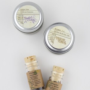 May include: Two silver tins with book titles and authors printed on the labels. The tins are labeled "Pride and Prejudice" by Jane Austen and "Catcher in the Rye, The" by J.D. Salinger. Two small glass bottles with cork tops contain loose leaf tea blends. The bottles are labeled "Fireside Evenings" and "Lavender Mint Dream".