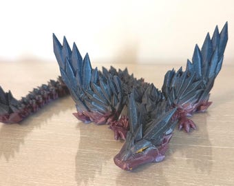 May include: A detailed figurine of a dragon with a crystal-like appearance. The dragon is primarily dark blue with red accents, and its body is covered in sharp, pointed structures. The dragon is lying down on a wooden surface.