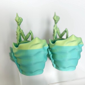 May include: Two 3D-printed teal and lime green planters, each with a small, light green figure seated inside. The planters have a layered, textured design. The figures have a humanoid shape with a pointed head.
